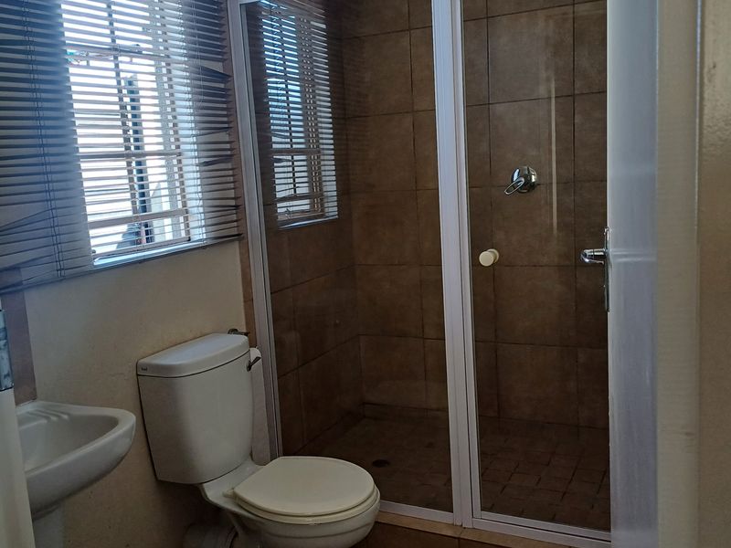 A functional bathroom featuring a shower enclosure, pedestal basin, and toilet with neutral wall tiling and natural light from the window.