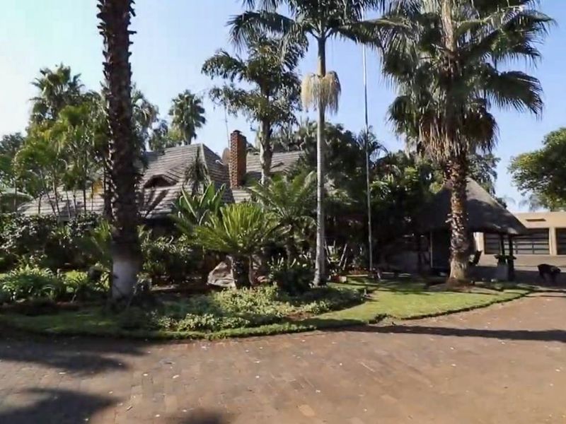 Front view of a property featuring a thatched roof house, brick chimney, mature landscaped garden, and a paved driveway leading to a garage.