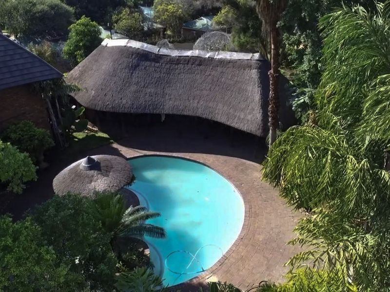 Aerial view of a swimming pool area featuring a large thatched lapa, paved surround, and established garden landscaping.