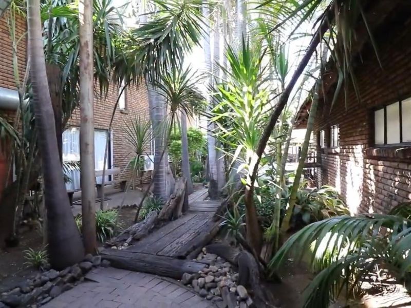 A lush, landscaped garden area featuring a timber walkway, paved paths, and established trees nestled between brick building structures.