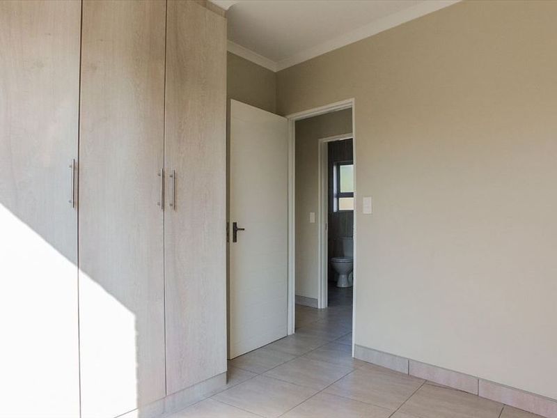 A clean, empty bedroom featuring light wood-finish built-in cupboards, neutral wall tones, and tiled flooring, with an adjacent bathroom visible through the doorway.