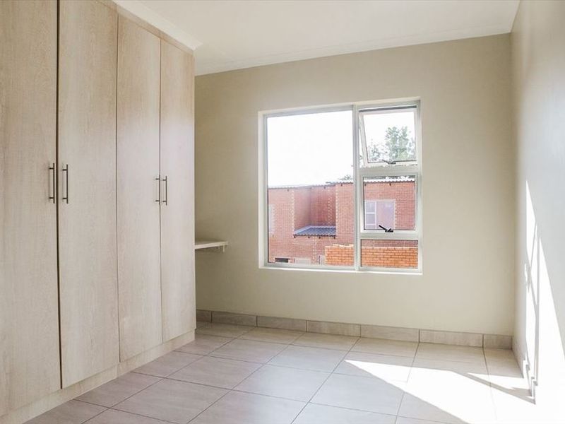 A bright, empty bedroom featuring neutral tiled flooring, built-in wooden cupboards, and a large window providing natural light.