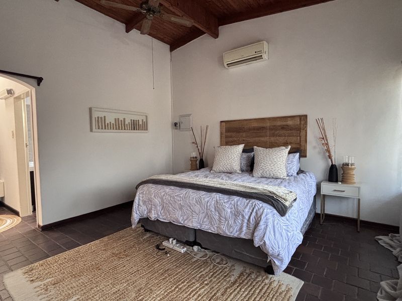 Spacious bedroom featuring exposed timber ceiling beams, tiled flooring, and a split-system air conditioner, with an adjoining en-suite bathroom area.