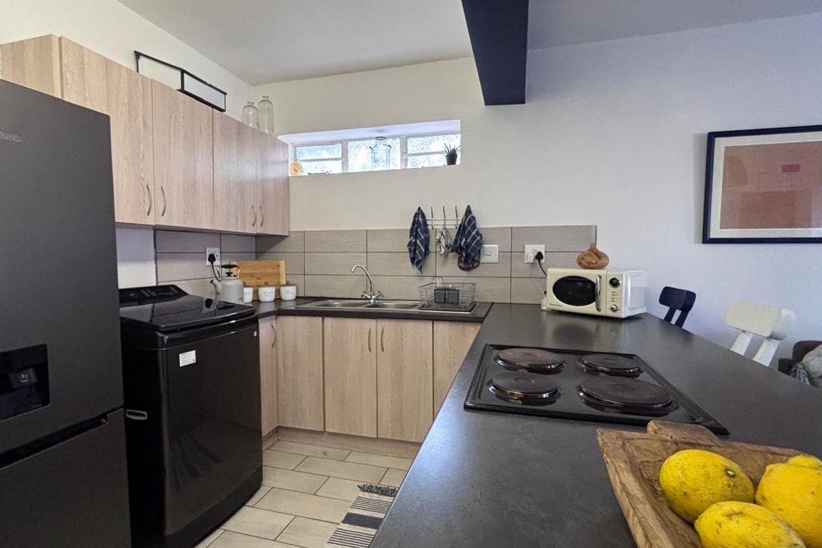 A functional kitchen featuring light wood-tone cabinetry, a dark countertop with an integrated electric hob, tiled backsplash, and a double sink.