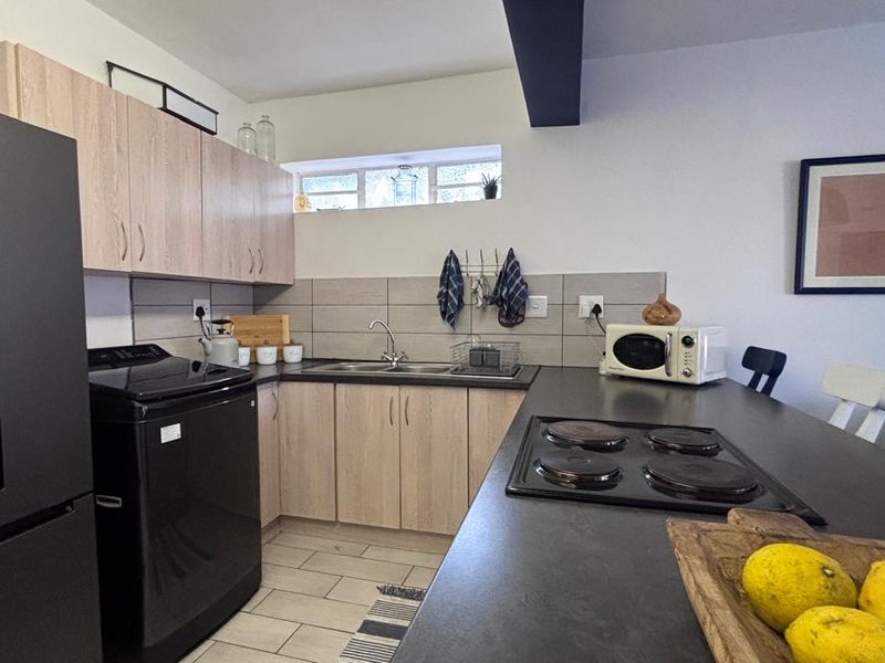 A functional kitchen featuring light wood-tone cabinetry, a dark countertop with an integrated electric hob, tiled backsplash, and a double sink.