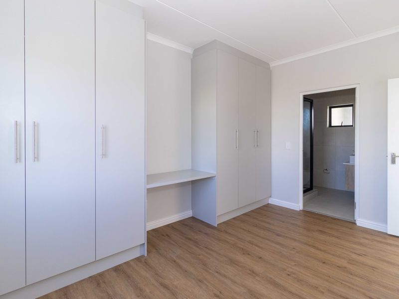 A bright, modern bedroom featuring neutral walls, laminate flooring, extensive built-in cabinetry with an integrated desk, and direct access to an en-suite bathroom.