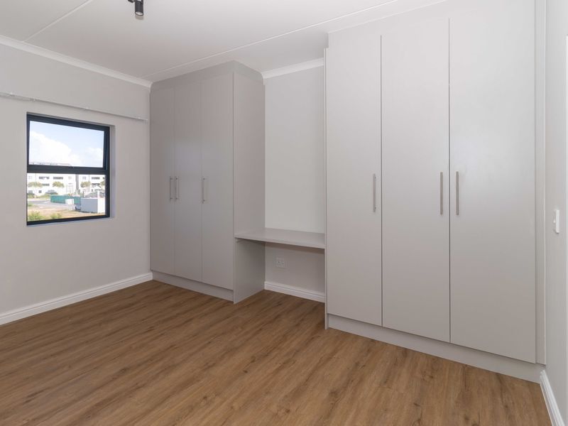 A bright, modern bedroom featuring neutral walls, wood-look laminate flooring, and extensive built-in cabinetry with an integrated desk space.