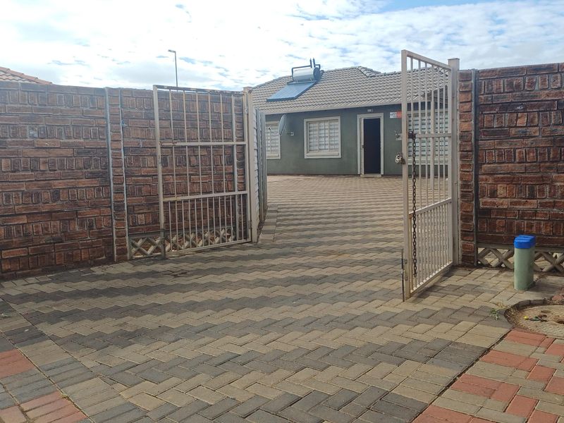 A secure residential property entrance featuring a paved driveway, brick boundary walls, metal security gates, and a house equipped with a solar water heater.