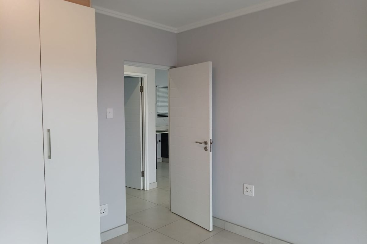 A clean, empty bedroom featuring neutral grey walls, tiled flooring, and a built-in wardrobe, with a doorway leading to an adjacent room.