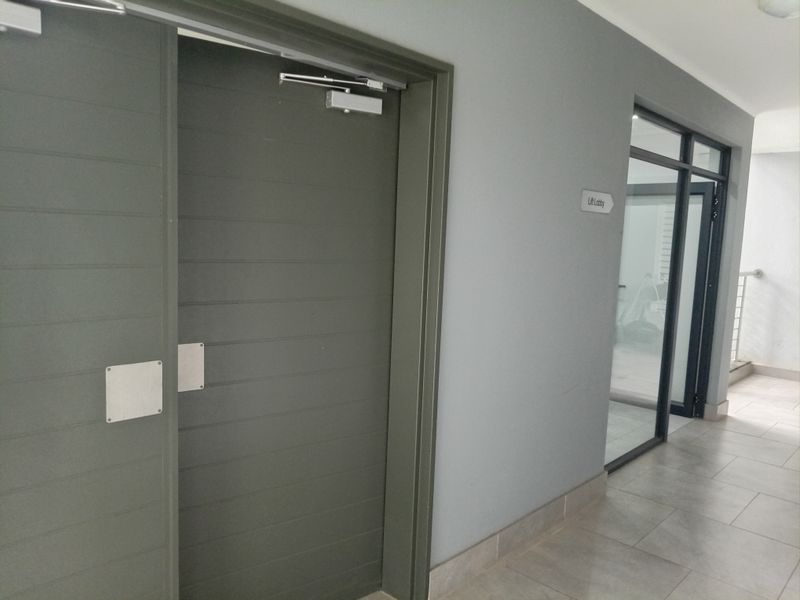A clean, modern apartment hallway featuring tiled flooring, a secure fire door with a closer, and a glass partition leading to the lift lobby.