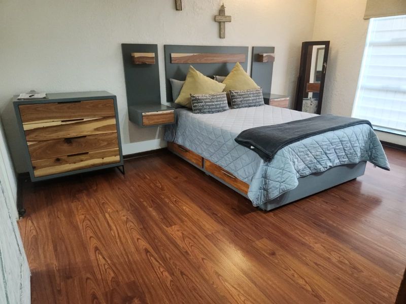A well-maintained bedroom featuring warm-toned laminate flooring, a custom built-in headboard with integrated floating nightstands, and a large window with a roller blind.