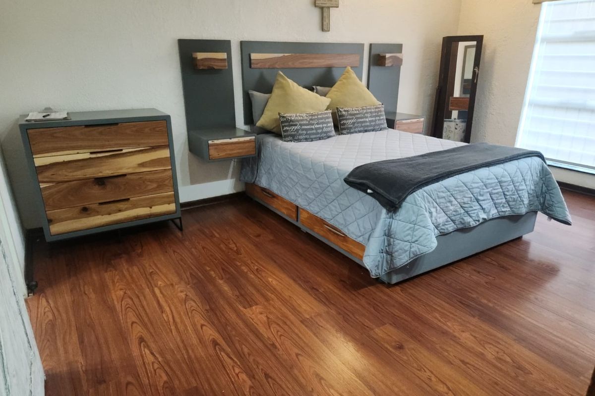 A well-maintained bedroom featuring warm-toned laminate flooring, a custom built-in headboard with integrated floating nightstands, and a large window with a roller blind.