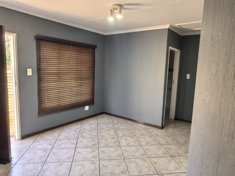A bright, empty living room featuring neutral grey walls, tiled flooring, and venetian blinds, offering a clean and versatile space for personalization.