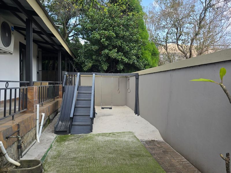 A well-maintained rear garden area featuring a covered patio, artificial lawn, paved pathways, and a perimeter wall with a built-in play structure.