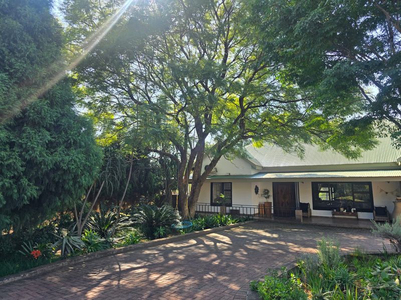 A charming front exterior view featuring a paved driveway, a covered porch, and a well-established garden with mature trees.