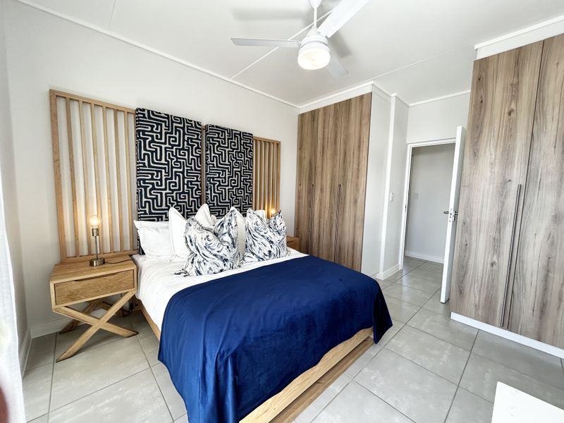 A modern bedroom featuring built-in wood-finish cupboards, tiled flooring, and a ceiling fan, presenting a clean and well-maintained living space.