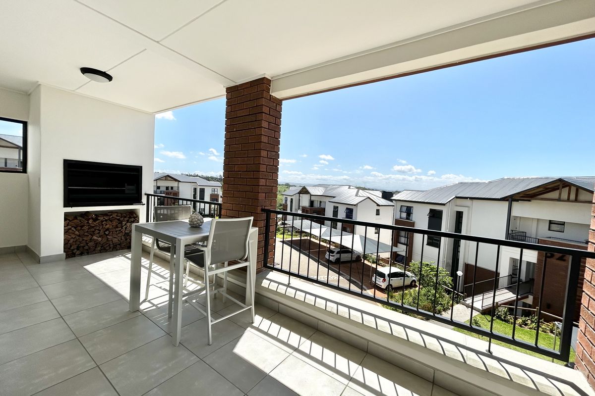 A spacious covered balcony featuring a built-in braai, tiled flooring, and a metal balustrade, offering pleasant views of the surrounding apartment complex.