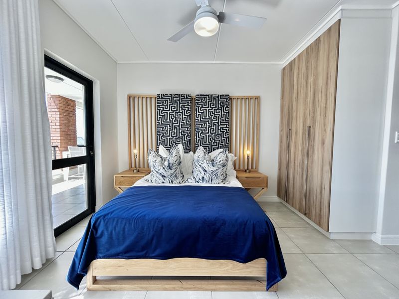 A well-maintained bedroom featuring built-in wooden cupboards, a ceiling fan, tiled flooring, and direct access to a balcony through a sliding glass door.