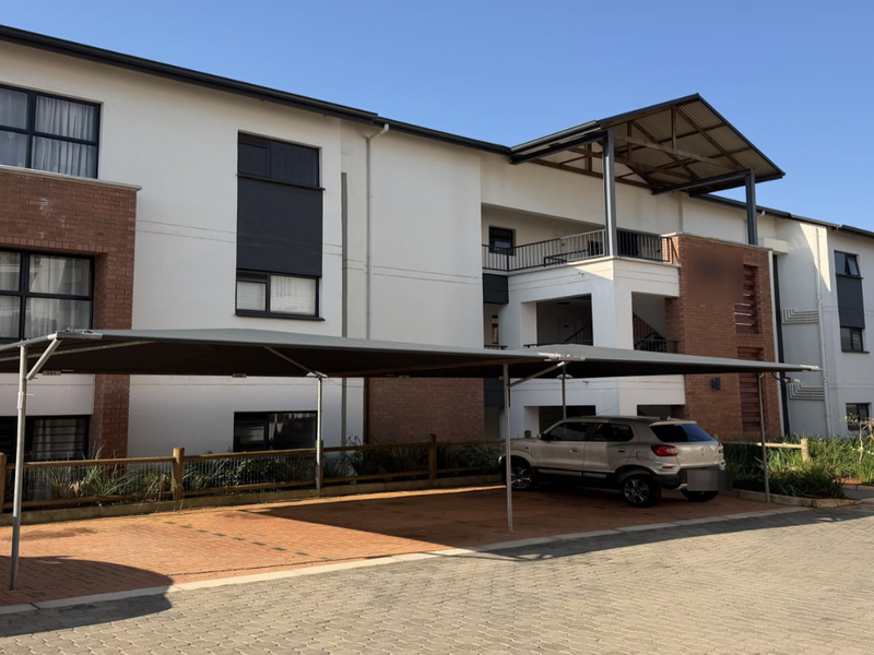 Exterior view of a modern apartment complex featuring a paved driveway, shade cloth carports, and brick-accented facade with balconies.