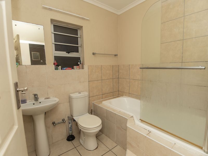 A functional bathroom featuring a pedestal basin, toilet, and a built-in bathtub with a glass shower screen, finished with neutral wall and floor tiles.