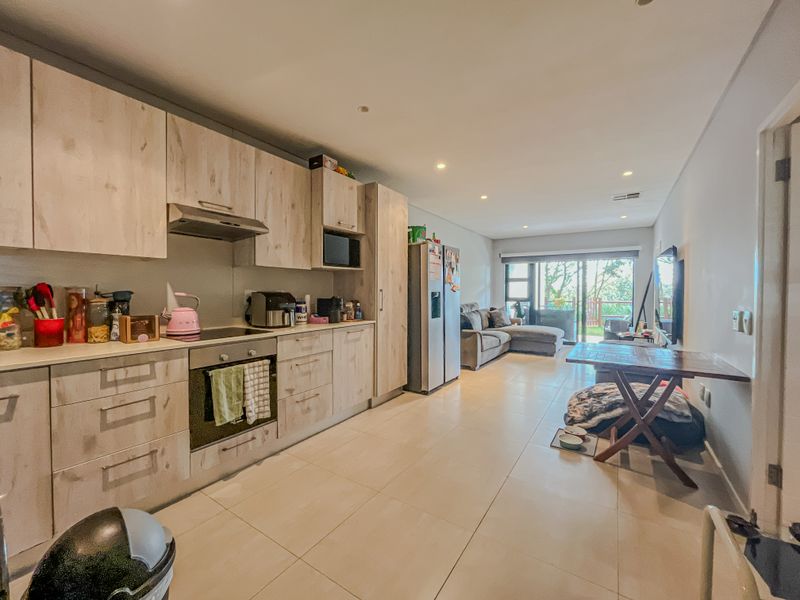 An open-plan living and kitchen area featuring light wood-finish cabinetry, modern appliances, tiled flooring, and direct access to an outdoor patio through sliding doors.