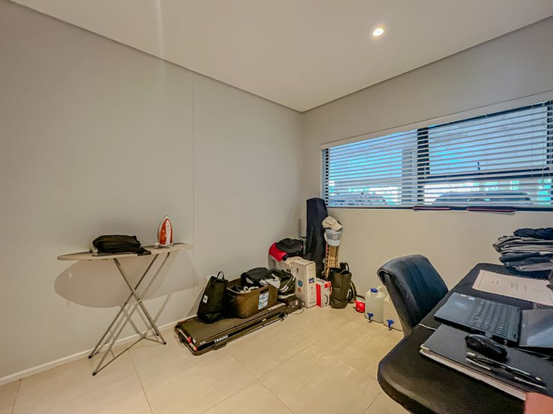 A functional office space featuring neutral tiled flooring, recessed downlighting, and a large window fitted with venetian blinds.