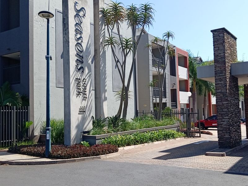 The entrance facade of Seasons Court Park apartment complex, featuring modern concrete signage, stone pillar detailing, and paved driveway access.