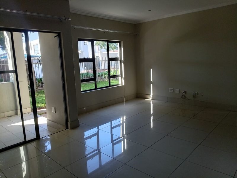 An empty, sunlit living room featuring polished tiled flooring, aluminium window frames, and a sliding glass door providing access to the exterior.