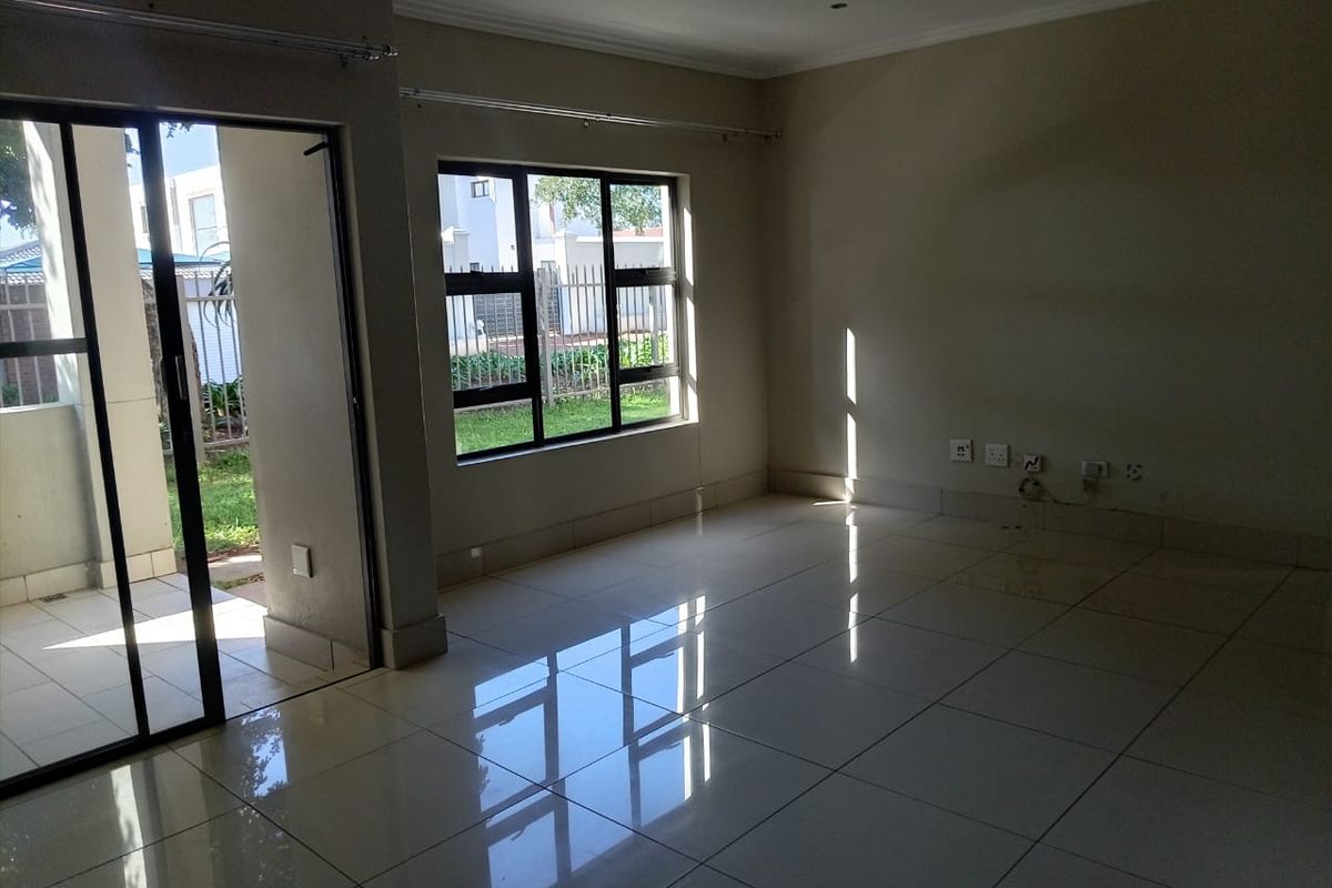 An empty, sunlit living room featuring polished tiled flooring, aluminium window frames, and a sliding glass door providing access to the exterior.