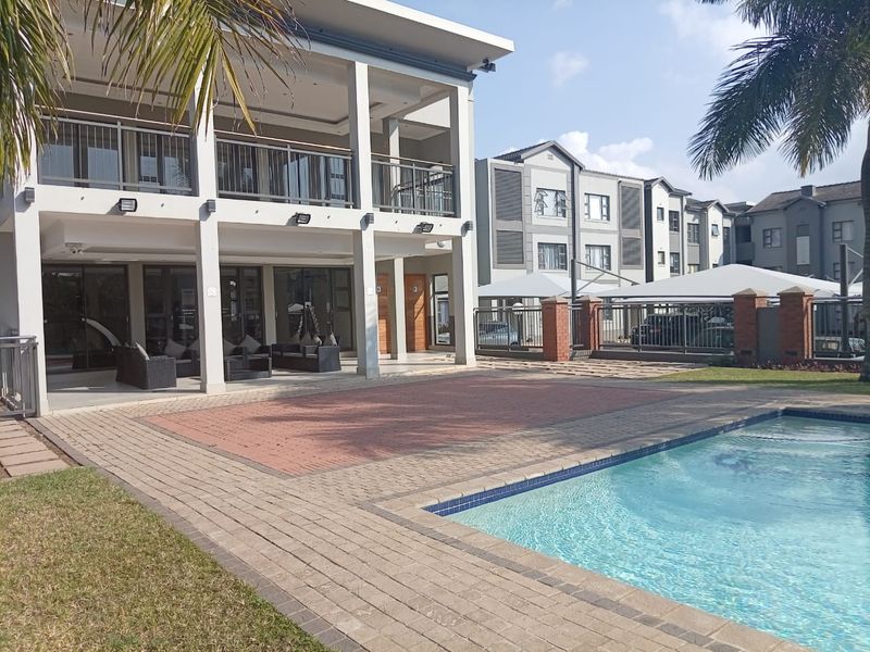 A well-maintained communal pool area featuring a paved deck, covered patio space, and multi-story apartment building architecture with balcony access.