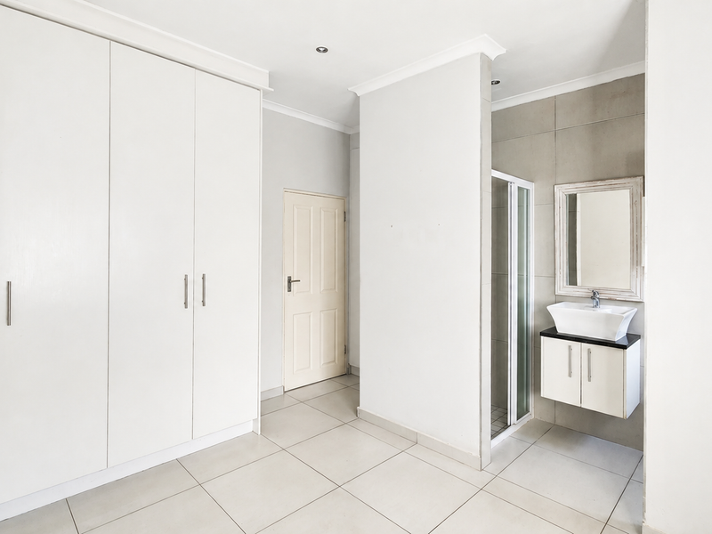 Spacious bedroom featuring built-in cupboards, neutral tiled flooring, and a convenient en-suite bathroom with a modern wall-mounted vanity and shower.
