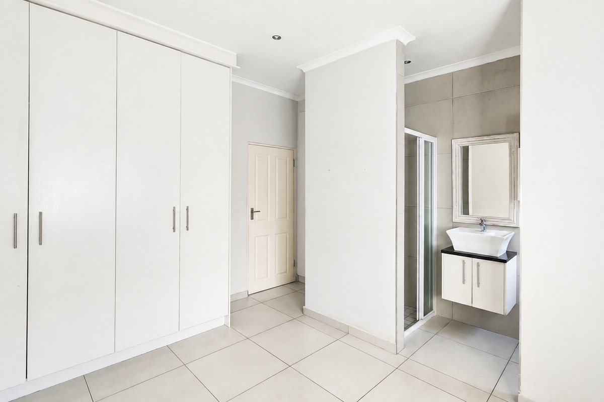 Spacious bedroom featuring built-in cupboards, neutral tiled flooring, and a convenient en-suite bathroom with a modern wall-mounted vanity and shower.
