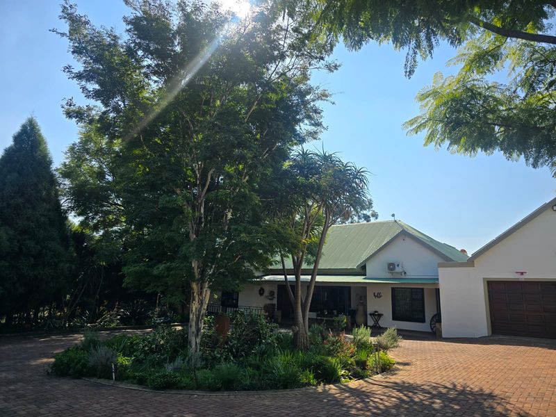 Front view of the property featuring a paved circular driveway, mature landscaped garden, and a well-maintained building facade with a metal roof.