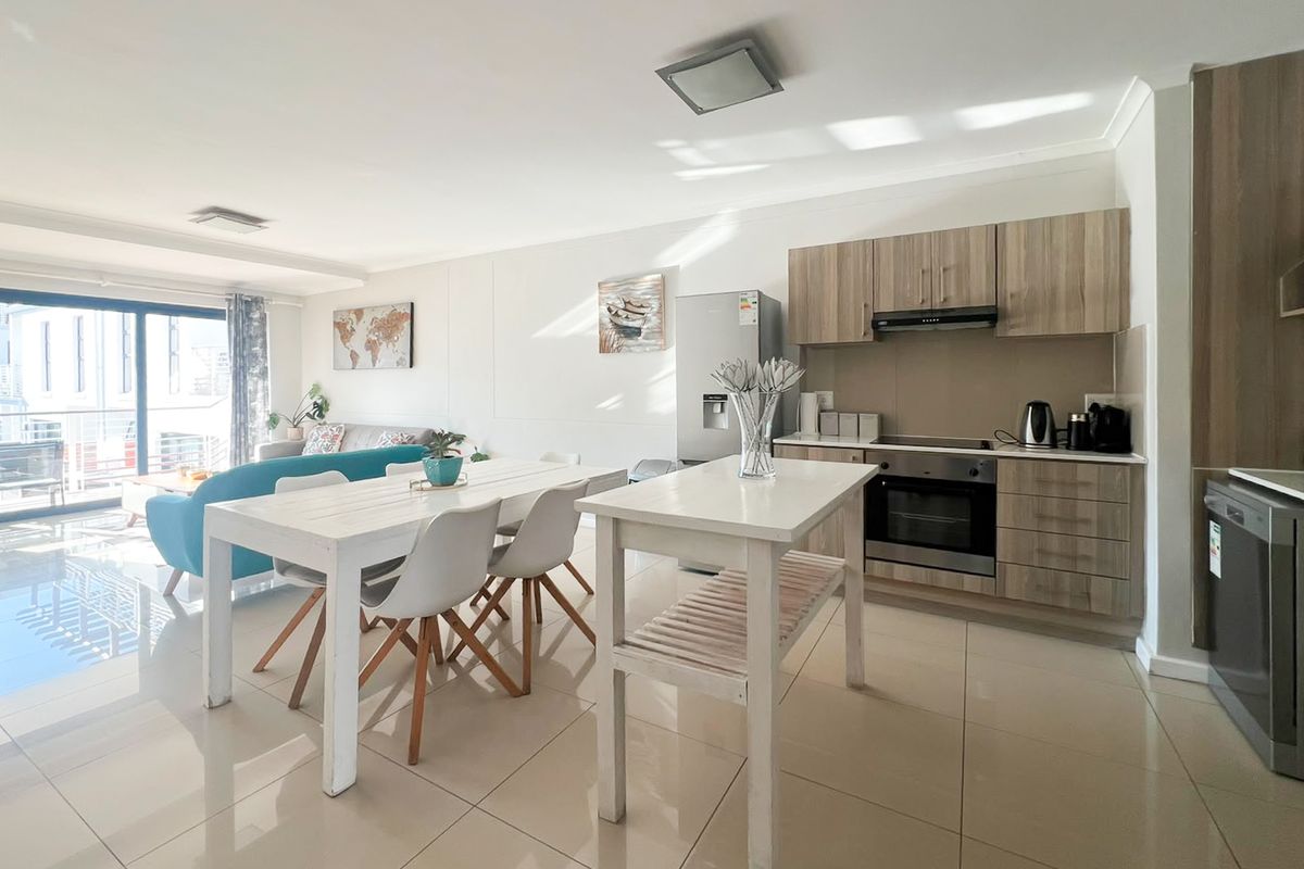 Modern open-plan living area featuring a contemporary kitchen with wood-finish cabinetry, integrated appliances, and seamless flow to a bright balcony through sliding glass doors.