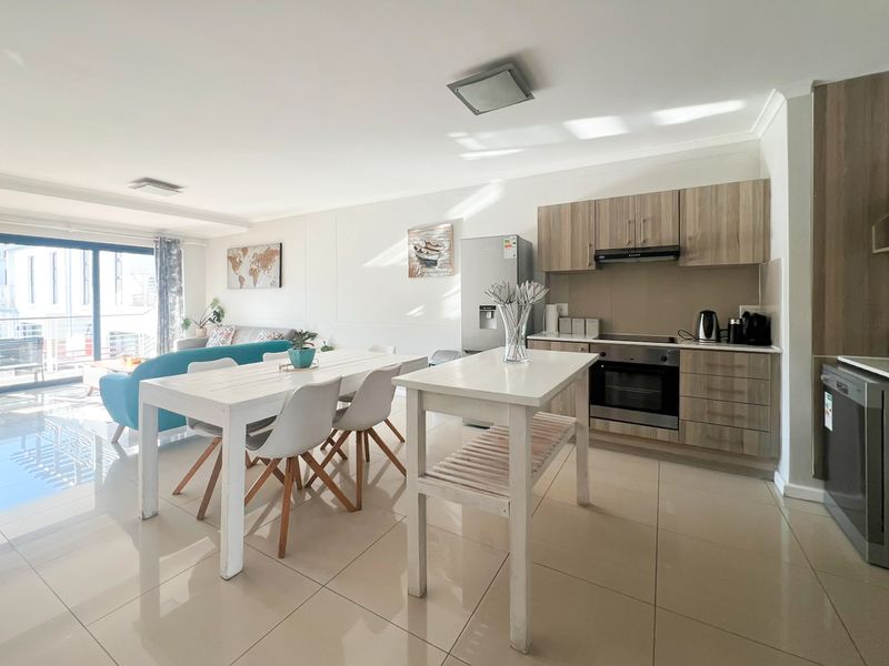 Modern open-plan living area featuring a contemporary kitchen with wood-finish cabinetry, integrated appliances, and seamless flow to a bright balcony through sliding glass doors.