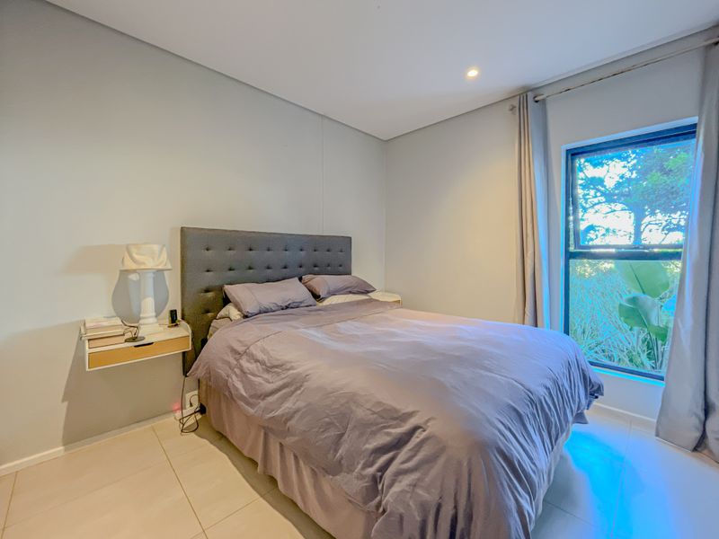 A well-maintained bedroom featuring neutral tiled flooring, recessed lighting, and a large window providing natural light and garden views.