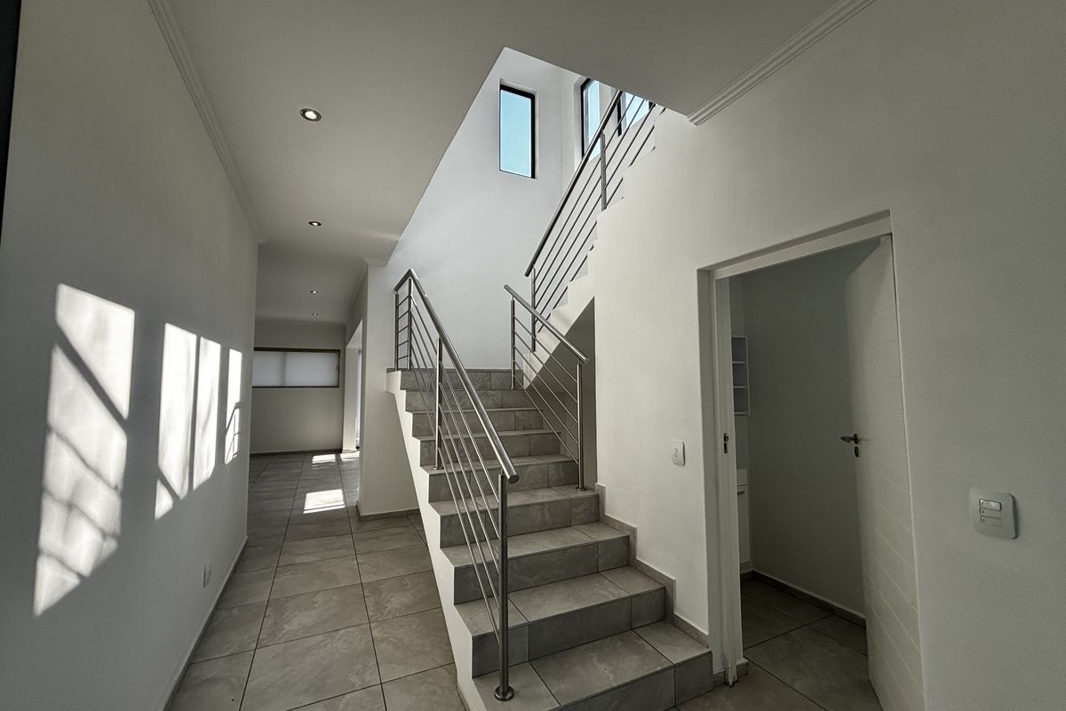 A bright, modern hallway featuring tiled flooring, a staircase with stainless steel railings, and recessed lighting, leading to an adjacent room.