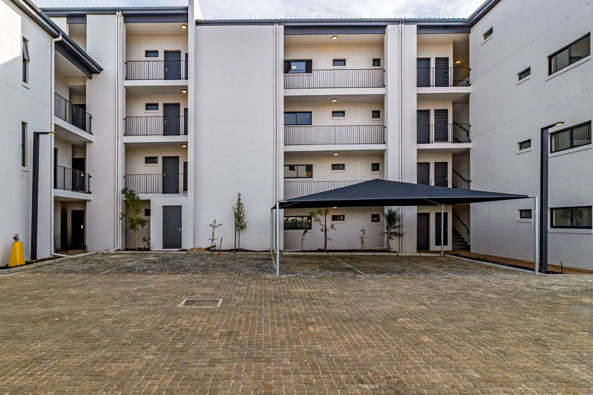 A modern apartment complex exterior featuring a paved courtyard, covered parking bays, and multi-level balconies with clean architectural lines.