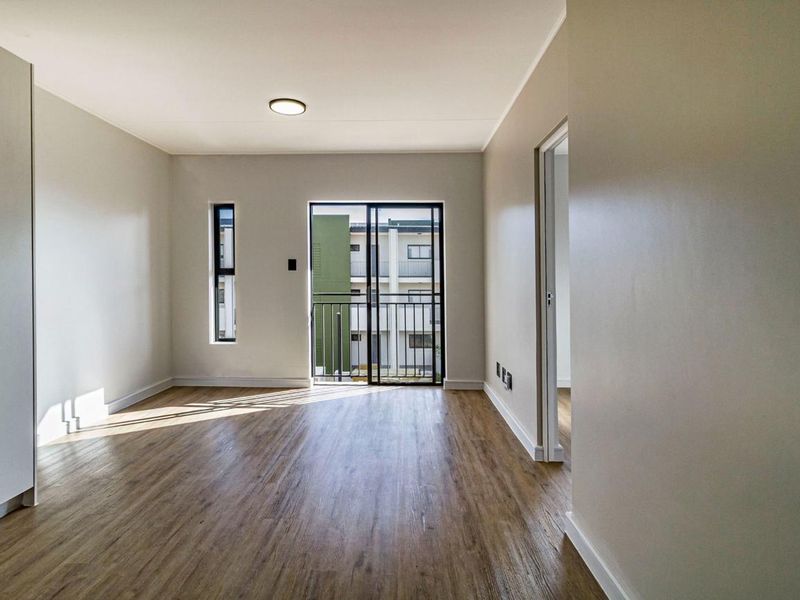 A bright, empty living room featuring modern laminate flooring, a sliding glass door leading to a balcony, and neutral wall finishes.