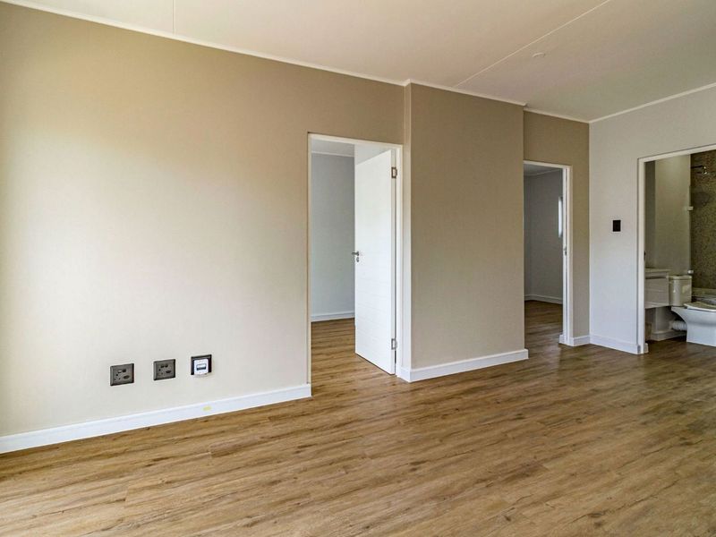 An empty, well-maintained living area featuring modern laminate flooring, neutral wall tones, and direct access to adjacent rooms and a bathroom.