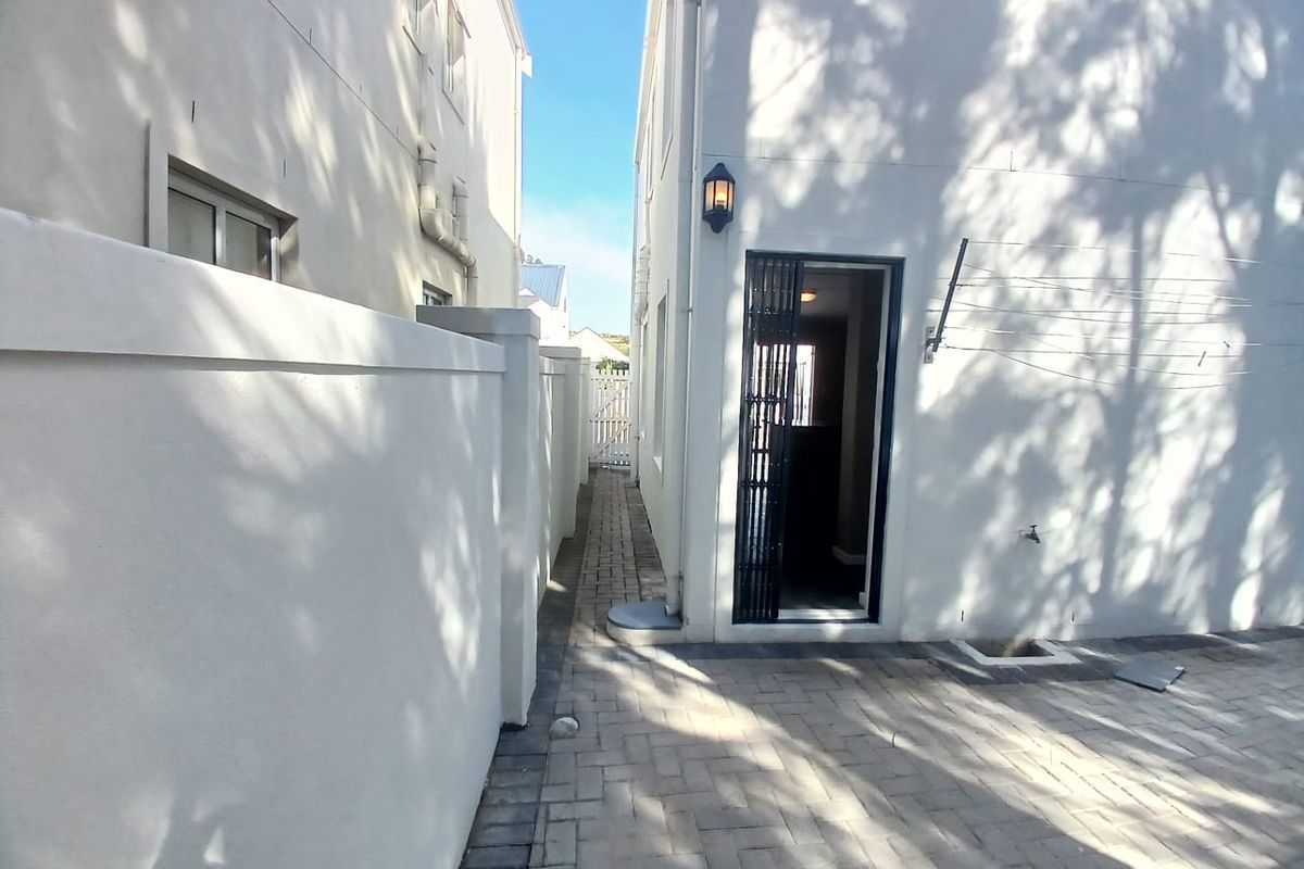 A clean, paved side passage and rear exterior area featuring a secure entrance gate, boundary wall, and functional outdoor lighting.