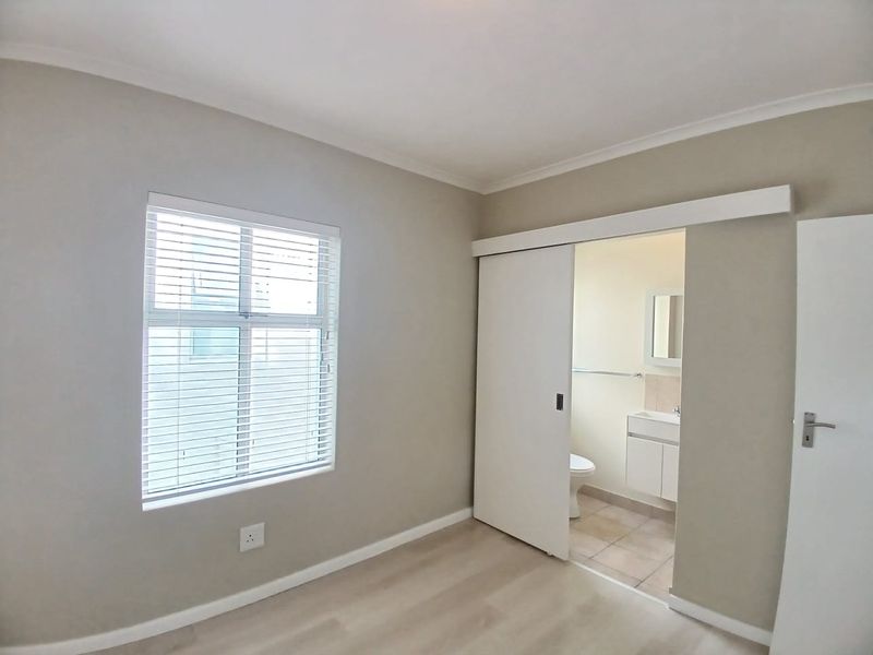 A clean, neutral-toned bedroom featuring laminate flooring, venetian blinds, and a sliding door leading to an en-suite bathroom with a vanity.