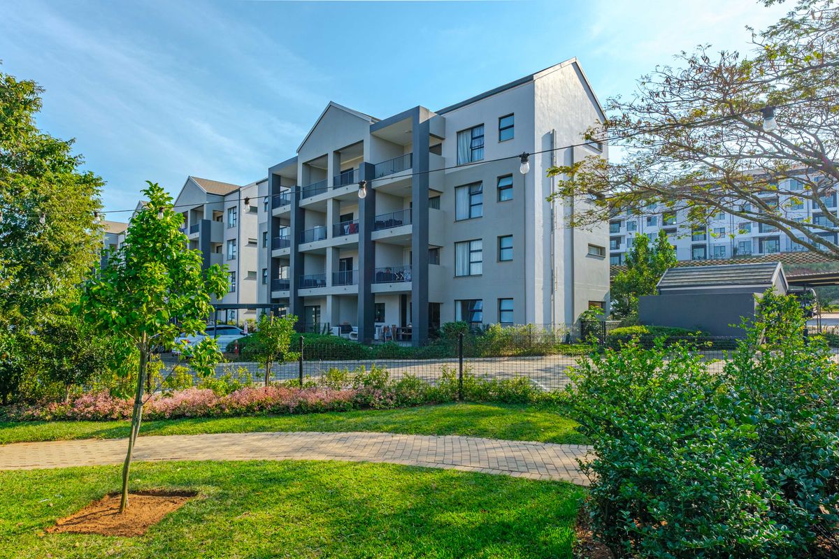 A view of the apartment complex exterior featuring multi-story balconies, a well-maintained garden, and a paved walkway.