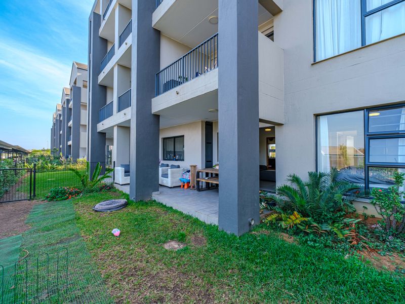 Ground floor apartment featuring a covered patio with tiled flooring, overlooking a private garden area with metal perimeter fencing and large sliding glass doors.