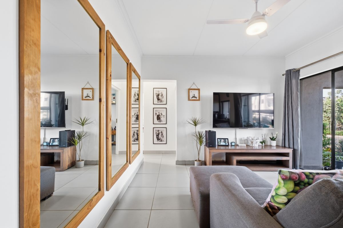 A bright living room featuring tiled flooring, a ceiling fan, and large wall-mounted mirrors, with direct access to an outdoor area via a sliding glass door.