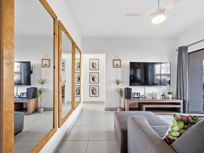 A bright living room featuring tiled flooring, a ceiling fan, and large wall-mounted mirrors, with direct access to an outdoor area via a sliding glass door.