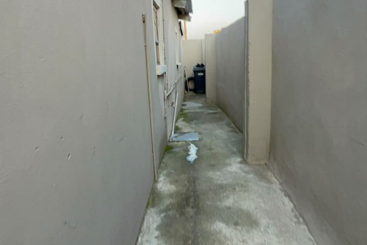A narrow side passage or rear exterior walkway featuring concrete paving and boundary walls, showing signs of weathering and surface staining.