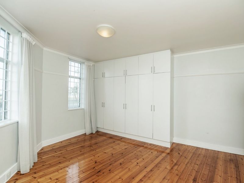 A bright, empty bedroom featuring polished timber flooring, built-in floor-to-ceiling white wardrobes, and large multi-pane windows.