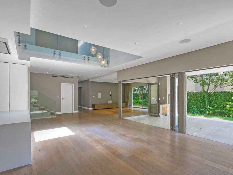Spacious open-plan living area featuring hardwood floors, recessed lighting, a glass balustrade, and seamless indoor-outdoor flow through stacking glass doors to the garden.