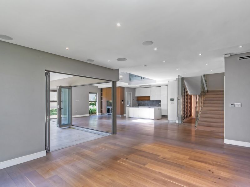 Spacious open-plan living area featuring hardwood floors, modern kitchen with island, glass-enclosed staircase, and seamless indoor-outdoor flow through folding glass doors.